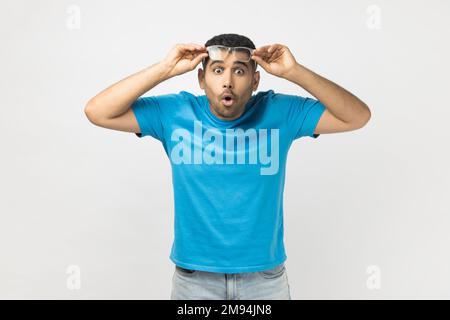 Portrait eines schockierten, erstaunten, unrasierten Mannes, der ein blaues T-Shirt trägt, seine optische Brille hochhob, sieht etwas Überraschtes. Studioaufnahmen im Innenbereich isoliert auf grauem Hintergrund. Stockfoto