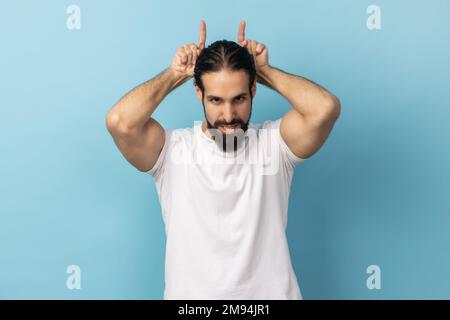 Porträt eines aggressiven Tyrannen mit Bart und weißem T-Shirt, mit Stierkopfgesten über dem Kopf, Stirnrunzeln wie vor dem Angriff, Indoor Studio-Aufnahme isoliert auf blauem Hintergrund. Stockfoto