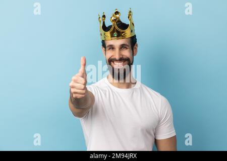 Porträt eines lächelnden, fröhlichen Mannes mit Bart, weißem T-Shirt und goldener Krone, der in die Kamera schaut, mit einem zahnigen Lächeln, mit dem Daumen nach oben zeigt. Innenstudio, isoliert auf blauem Hintergrund. Stockfoto
