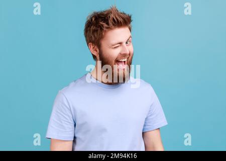 Porträt eines bärtigen Mannes, der gut gelaunt ist, weites Lächeln und vor der Kamera blinzelt, den Mund offen hält, mit flirtendem Gesichtsausdruck. Innenstudio, isoliert auf blauem Hintergrund. Stockfoto