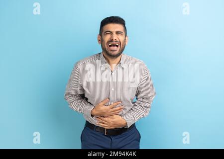 Porträt eines lustigen, gutaussehenden, jungen Geschäftsmannes, der mit geschlossenen Augen steht und lacht, seinen Bauch hält, ein gestreiftes Hemd trägt. Innenstudio, isoliert auf blauem Hintergrund. Stockfoto