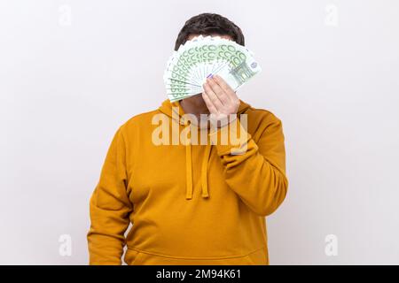 Ein unbekannter Mann versteckt sich hinter einem Fan von Hundert-Euro-Scheinen, gewinnt, wettet, trägt einen urbanen Hoodie. Studioaufnahmen im Innenbereich isoliert auf weißem Hintergrund. Stockfoto