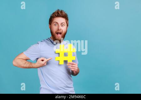 Portrait eines überraschten gutaussehenden Mannes, der mit dem Finger auf das Hashtag in seiner Hand zeigt, mit offenem Mund und großen Augen, Website-Suche. Innenstudio, isoliert auf blauem Hintergrund. Stockfoto