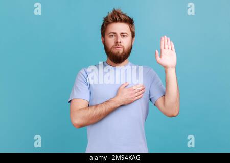 Portrait eines ehrlichen, bärtigen Mannes, der mit Hand auf Herz Versprechen gibt, Treue schwört, einen Eid mit verantwortungsvollem, ernstem Gesicht abgibt. Innenstudio, isoliert auf blauem Hintergrund. Stockfoto