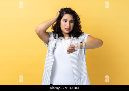 Porträt einer Frau mit dunklem, welligem Haar, die mit verwirrtem Gesichtsausdruck steht, sich Sorgen um die Frist macht, auf die Armbanduhr schaut, die Hand am Kopf hält. Innenstudio-Aufnahme isoliert auf gelbem Hintergrund. Stockfoto