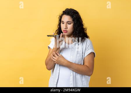 Digitale Lautsprecher-App. Porträt einer Frau mit dunklem, welligem Haar, die mit dem Mobiltelefon spricht, mit Sprachassistent Erinnerungen aufzeichnet, Nachrichten. Innenstudio-Aufnahme isoliert auf gelbem Hintergrund. Stockfoto