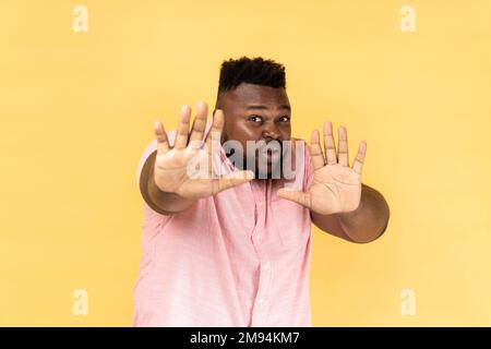 Nein, es ist beängstigend. Porträt von einem verängstigten, geschossenen Mann, der ein rosa Hemd trägt, die Hände hebt in Angst, entsetzt und panisch aussieht, sich vor Phobie versteckt. Innenstudio-Aufnahme isoliert auf gelbem Hintergrund. Stockfoto