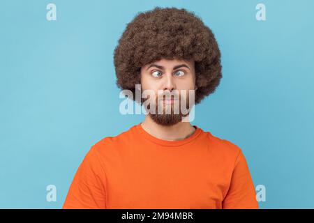 Portrait eines verrückten, lustigen Mannes mit Afro-Haarschnitt, der ein orangefarbenes T-Shirt trägt, mit gekreuzten Augen steht und mit einem albernen komischen Gesicht aussieht. Innenstudio, isoliert auf blauem Hintergrund. Stockfoto