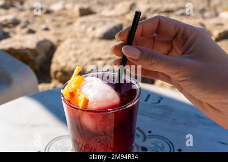 Von oben, anonyme weibliche Hand, die an sonnigen Tagen einen roten Cocktail in Glas mit Stroh rührt Stockfoto