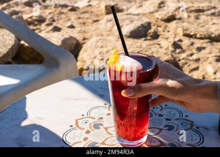 Eine anonyme, aus einem hohen Winkel gefertigte Frau nimmt ein Glas erfrischenden roten Cocktail mit Eiswürfeln und Stroh, während sie sich in der Cafeteria im Freien ausruht Stockfoto