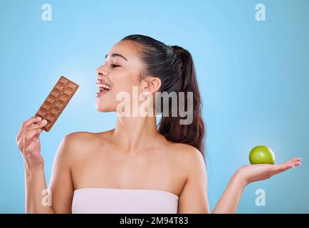 Sie hat ihre Wahl getroffen. Studio hat eine attraktive junge Frau erschossen, die sich vor blauem Hintergrund zwischen Schokolade und Apfel entschieden hat. Stockfoto