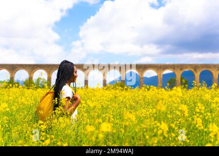 Genießen Sie den Frühling, schwarzes ethnisches Mädchen mit Zöpfen, Reisende, auf einem Feld gelber Blumen Stockfoto