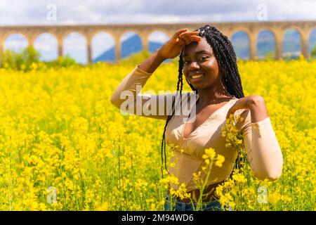 Lifestyle, Porträt eines schwarzen ethnischen Mädchens mit Zöpfen, das in die Kamera schaut, Trap-Musiktänzerin, in einem Feld gelber Blumen Stockfoto