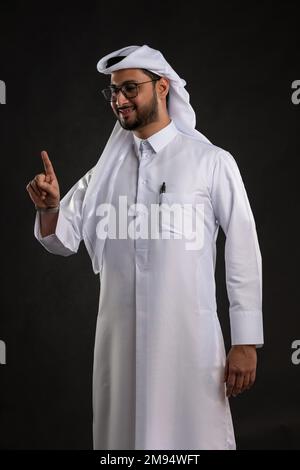 Arabischer Katar-Mann, der Tisch benutzt und auf isoliertem Hintergrund posiert. Männliches Modell in arabisch-katarischem Kleid auf isoliertem Hintergrund. Verschiedene Posen Stockfoto