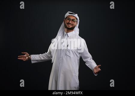 Arabischer Katar-Mann, der Tisch benutzt und auf isoliertem Hintergrund posiert. Männliches Modell in arabisch-katarischem Kleid auf isoliertem Hintergrund. Verschiedene Posen Stockfoto