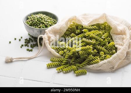 Grüne Mungbohnen-Fusilli-Pasta auf weißem Fliesenhintergrund. Beutel mit roher Pasta und Schüssel mit Mungbohne. Glutenfreie Nudeln. Stockfoto