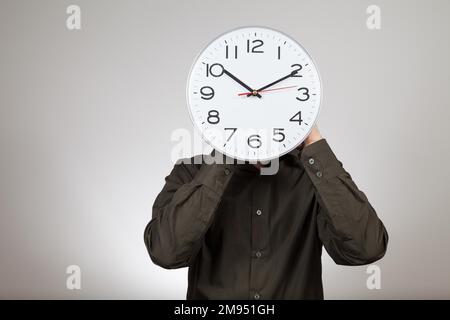 Symbolisches Bild der Arbeitszeit, Mann hält eine Uhr vor seinem Gesicht Stockfoto