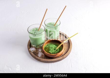 eistee Matcha dalgona in zwei Gläsern mit einem Strohhalm und einer Schüssel Matcha-Pulver zum Kochen auf einem Holztablett. Weißer Hintergrund. Platz für Text. Stockfoto
