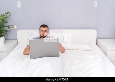 Ein Mann mit Laptop liegt im Bett. Freiberufler arbeiten von zu Hause aus. Fernunterricht. Online-Shopping ohne das Haus zu verlassen. Surfen im Internet. Ga Stockfoto