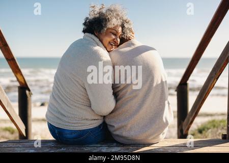 Rückblick auf eine glückliche ältere Frau, die fröhlich lächelt, während sie mit ihrem Mann vor dem Meer sitzt. Rentnerpaar, das Qualität verbringt Stockfoto