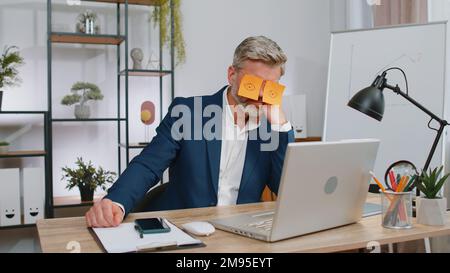 Ineffizienter, ermüdeter Geschäftsmann, der auf einem Laptop mit Augenaufklebern auf dem Gesicht am Schreibtisch im Büro schläft. Ein fauler Manager-Freiberufler mittleren Alters. Geschäftsleute schummeln in den Schlaf Stockfoto