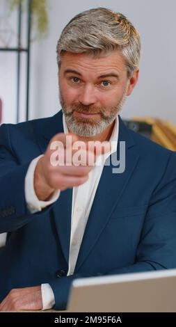 Ich wähle dich. Programmierer im mittleren Alter, der an einem Büro-Laptop arbeitet und auf die Kamera zeigt, mit fröhlichem Ausdruck aussieht, eine Wahl trifft, die Richtung zeigt. Reifer Freiberufler Stockfoto