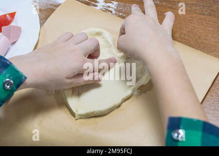 Auf einem hölzernen Küchentisch bilden die vom Kind geschnittenen Hände Teig zu einer kleinen herzförmigen Pizza. Stockfoto