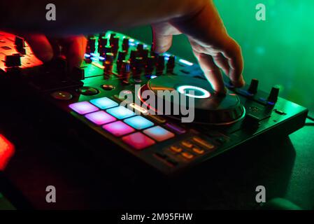 Mixer und DJ-Stand im Nachtclub, wobei die Arme des DJs die Musik mit bunter Beleuchtung und einem unscharfen grünen Hintergrund steuern Stockfoto