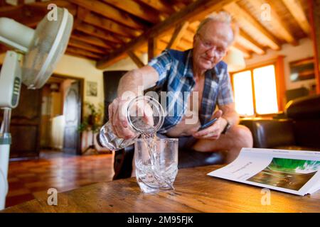 Senioren und Hitzewelle: Immer hydriert. Intensive Hitze: Alter Mann trinkt Wasser und Ventilator bläst Luft. Illustration, ältere Menschen und Hydratation, Stockfoto