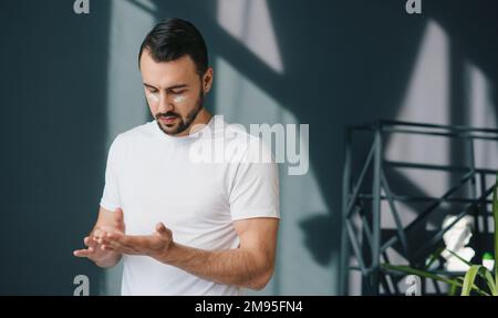 Ein junger Mann, der morgens im Wohnzimmer stand und sich mit Sahne aufs Gesicht legt, tägliche Routine. Natürliche Schönheit. Positive Person. Schönheitsgesicht. Körper positiv Stockfoto