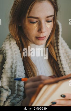Nahaufnahme Porträt einer Frau mit roten Haaren und Sommersprossen, die mit Bleistift und Notizbuch zu Hause in Quarantäne schreibt. Bleiben Sie zu Hause. Stockfoto