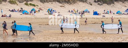 Ein Panoramabild von Urlaubern, die nach einem Surfunterricht am Fistral Beach in Newquay in Cornwall, Großbritannien, ihre Surfbretter tragen. Stockfoto