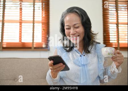 Glückliche Asiatin im Alter von 60s Jahren, die lacht, während sie ihr Smartphone benutzt, mit ihren Freunden chattet oder lustige Videos in sozialen Medien ansieht, während sie danach Spaß hat Stockfoto