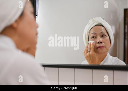 Eine schöne, alte Asiatin, die ihr Gesicht mit einem Wattepad reinigt und in den Spiegel schaut, nach dem Duschen Hautpflege macht. Stockfoto