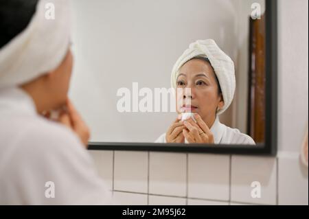 Wunderschöne 60s-jährige Asiatin, die ihr Make-up mit Make-up-Entferner und Wattepad entfernt, in den Spiegel schaut, sich auf die Dusche vorbereitet. Stockfoto