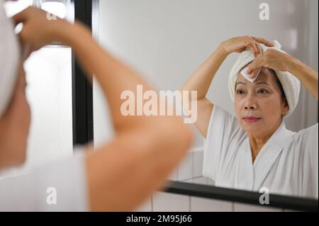 Schöne reife asiatische Frau reinigt die Gesichtshaut mit Reinigungsmittel, entfernt Make-up mit Reinigungsöl und Wattepad, schaut in den Spiegel. Schönheit Stockfoto