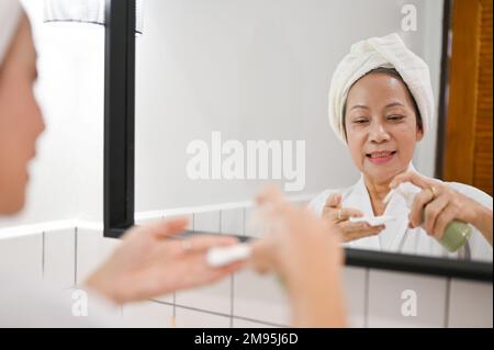 Eine attraktive, reife Asiatin steht im Badezimmer vor dem Spiegel, trägt Reinigungsöl oder Make-up-Entferner auf ein Wattepad auf, entfernt Gesicht m. Stockfoto