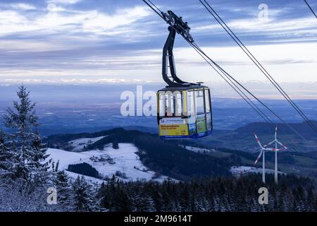 Freiburg, Deutschland. 17. Januar 2023. Ein Passagier fährt in einer Gondel der Schauinsland-Seilbahn hinunter in das Tal, während zwei Windturbinen und das schneefreie Rheintal im Hintergrund zu sehen sind. In den letzten Tagen waren mehrere Zentimeter Schnee gefallen, und Meteorologen zufolge werden in den nächsten Tagen weitere Zentimeter Schnee erwartet. Kredit: Philipp von Ditfurth/dpa/Alamy Live News Stockfoto