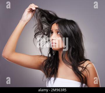 Warum ist es so verknotet? Eine attraktive junge Frau, die allein im Studio steht und sich gestresst fühlt, während sie ihr Haar ansieht. Stockfoto