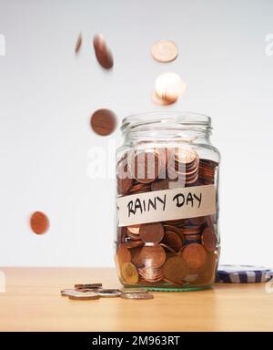 Ein altes Glasgefäß gefüllt mit Ersparnissen von vielen Münzen auf einem Holztisch. Einige Münzen fallen auf den Tisch. Stockfoto
