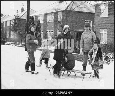 Mütter und Kinder, die mit einem Schlitten im Schnee in einer Vorstadtstraße in Horley, Surrey, England, spielen. Stockfoto