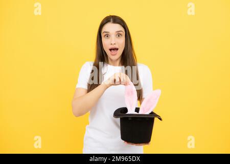 Kaninchenohren erscheinen aus dem magischen Hut. Hübsches Mädchen mit Hasenohren isoliert auf gelbem Hintergrund. Stockfoto