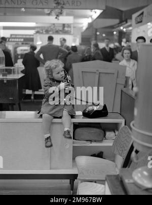 Ein kleines Mädchen am Telefon, das auf einem Stand auf einer Messe sitzt. Der Stand des „Midlands Gas Board“ ist im Hintergrund zu sehen. Foto von Norman Synge Waller Budd Stockfoto