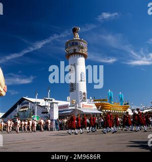 Szene auf dem Oktoberfest in München, Deutschland, mit einer Marschkapelle in Volkskleidern, die am Turm der Lowenbrau Brauerei vorbeizieht. Stockfoto