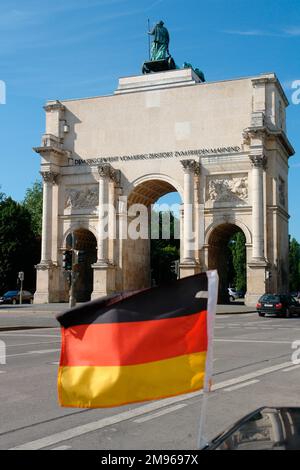 Blick auf eine winkende deutsche Flagge vor dem dreifach gewölbten Siegestor (Siegestor oder Triumphbogen) in München, Bayern. Das Tor wurde 1852 fertiggestellt und der bayerischen Armee gewidmet. Nach schweren Schäden während des Zweiten Weltkriegs wurde es restauriert und ist jetzt ein Friedensdenkmal. Stockfoto