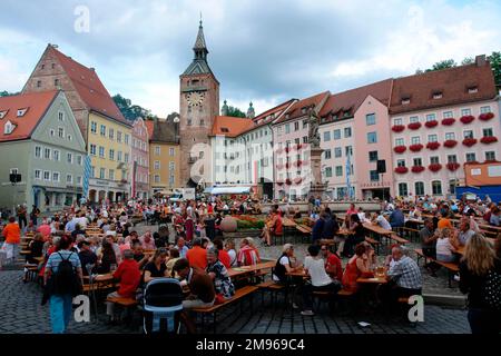 Wochenendfestival auf dem malerischen Marktplatz Landsberg am Lech, Bayern, mit Menschen, die an Böschungstischen sitzen. Der Schönestürm (wunderschöner Turm) neben dem alten Rathaus ist links vom Zentrum im Hintergrund zu sehen. Stockfoto