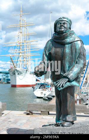 Statue von Evert Axel Taube, schwedischer Schriftsteller, Künstler, Komponist und Sänger (1890-1976) mit dem viermagigen Barbecue „Wikinger“ im Hintergrund, in Göteborg (Göteborg), Vastergotland, Schweden. Taube wurde in Göteborg geboren und wuchs auf der Insel Vinga auf, wo sein Vater, ein Schiffskapitän, der Leuchtturmwärter war. Taube ist bekannt für seine Volkslieder. Stockfoto