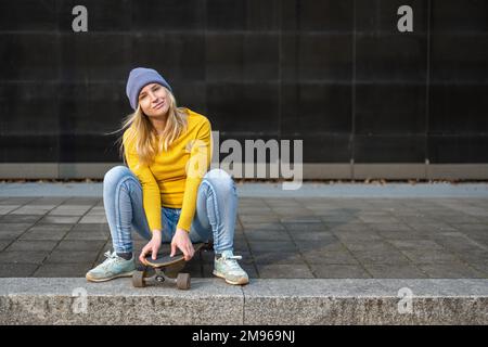 Junge Teenagerin in gelbem Trikot und Jeans, sitzt auf einem Skateboard, schaut in die Kamera und lächelt, Lifestyle der Z-Generation Stockfoto