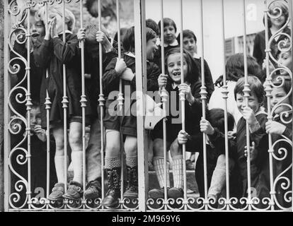 Eine glückliche Gruppe von Kindern, die durch ihre Schultore schauen, wahrscheinlich aufgeregt, fotografiert zu werden. Ihr Lehrer lächelt im Hintergrund rechts. Stockfoto