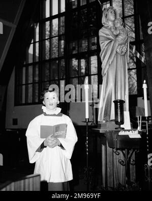 Ein Chorknabe, der allein vor einem modernen Buntglasfenster in der Salfords Church, Horley, Surrey singt. Er hält ein Buch mit dem Titel "englisches Lob". Neben ihm steht eine Statue der Jungfrau Maria und des Kindes. Stockfoto